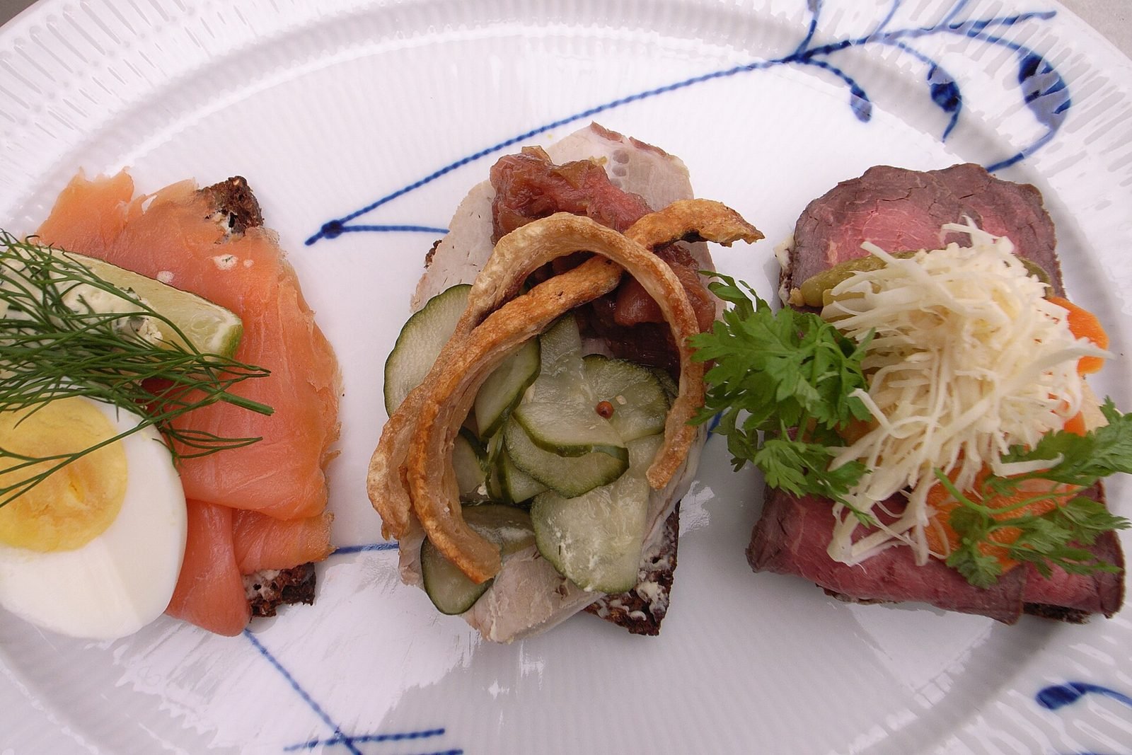 Drei belegte Sm&oslash;rrebr&oslash;d-Scheiben mit Lachs und Ei, Hering und Roastbeef auf einem blau gemusterten Teller